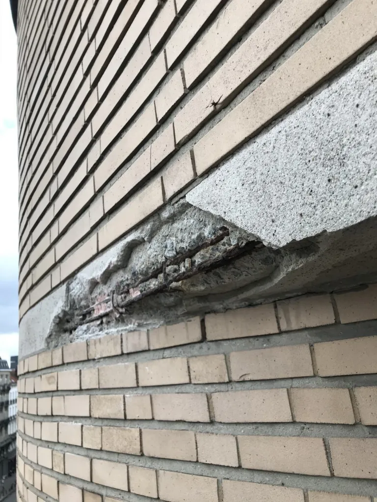 Un bandeau de façade en béton éclaté, armatures rouillées apparentes sous les briques