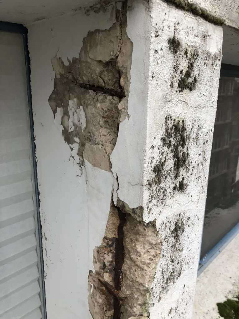 Un montant de façade en béton blanc éclaté, armatures rouillées apparentes, traces noires