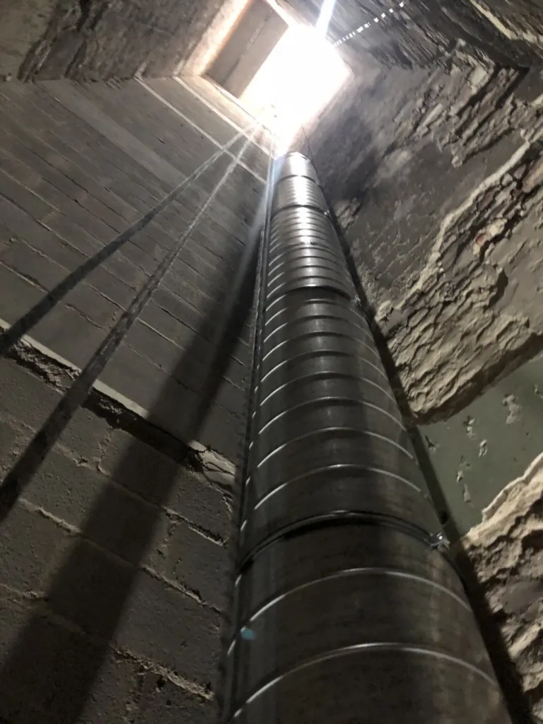 Un conduit métallique spiralé monte le long d'une gaine en brique jusqu'à l'ouverture en toiture