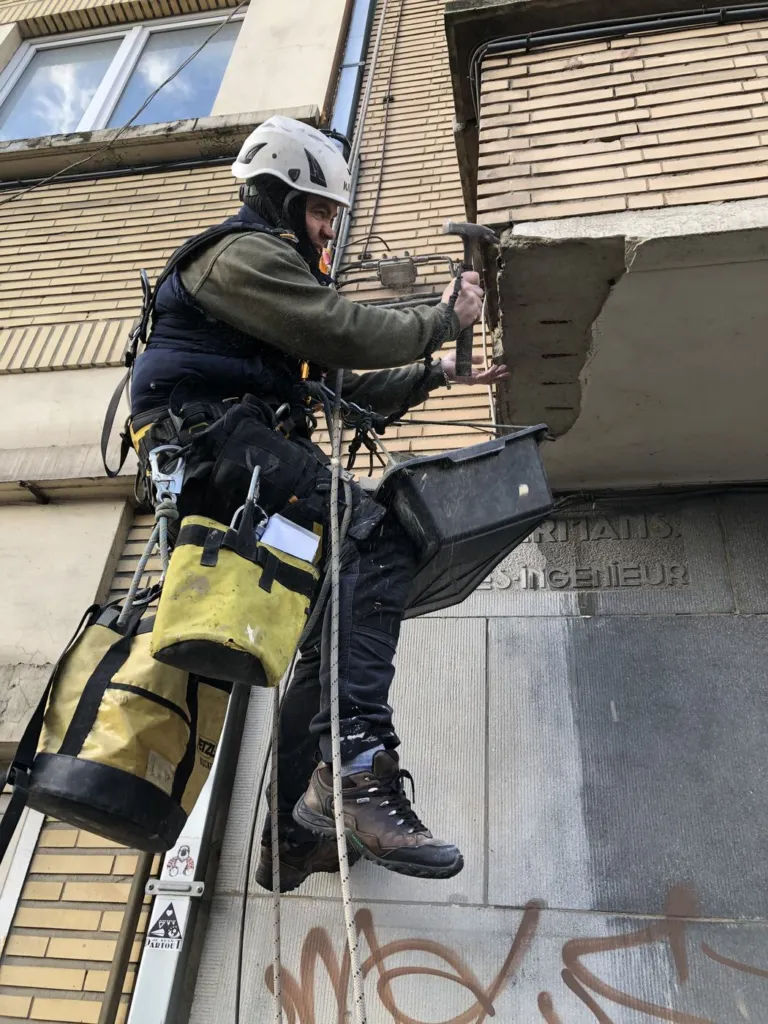 Un cordiste purge au marteau un bandeau de béton éclaté, bac de récupération accroché au harnais