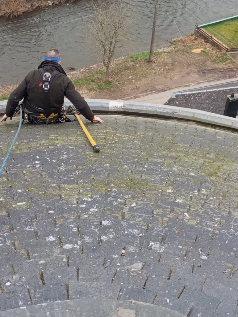 Un cordiste sur une couverture en ardoise couverte de mousse, l'outil à long manche en main, la rivière en contrebas