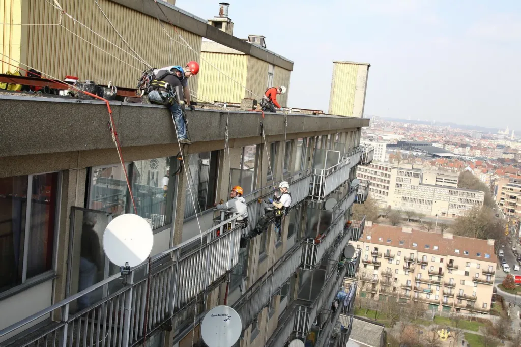 Plusieurs cordistes répartis sur la façade, vus depuis la toiture, la ville derrière