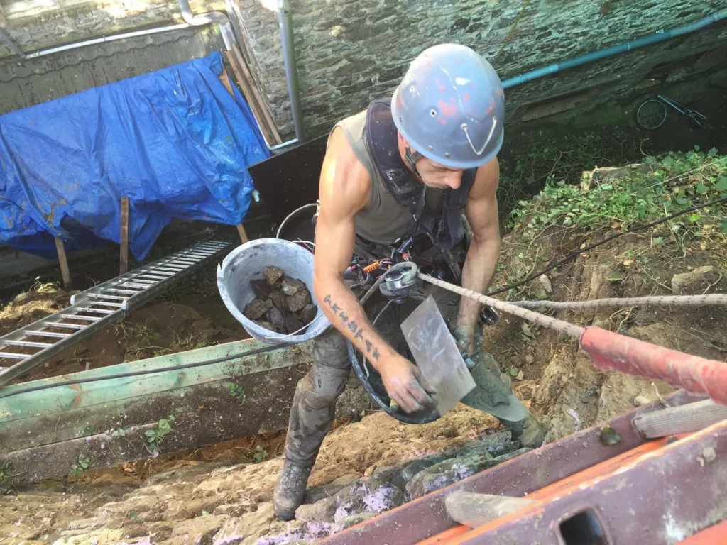 Un cordiste en harnais et casque, truelle en main et seau de mortier au bras, en train de maçonner