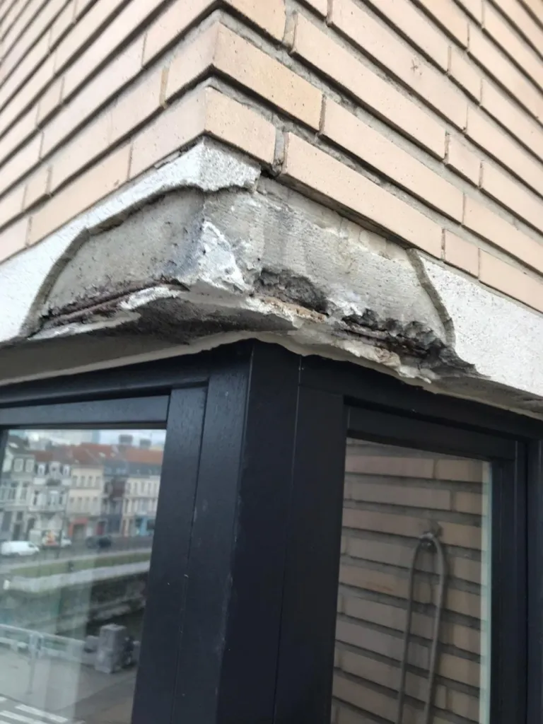 Le béton d'un angle de façade éclaté par son armature, sous le parement de briques