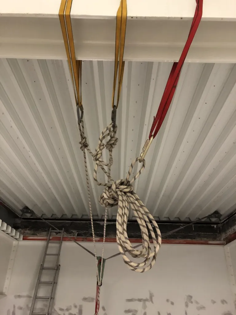 Sangles d'amarrage jaunes et rouge passées autour d'une poutre, cordes nouées et lovées en dessous, avant une descente en silo